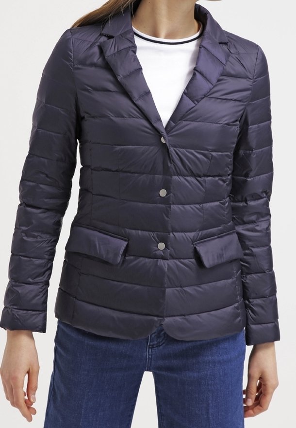 Kvinna i en marinblå kviltad kavaj med spetsiga lappar, knappstängning, fickor med klaff, över en vit t-shirt med rund halsringning och blå jeans.