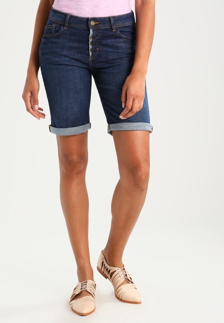 Bermudas de mezclilla en azul oscuro con dobladillos enrollados, que presentan un cierre de cinco botones. Combinadas con zapatos claros con cordones.