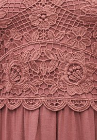 Λεπτομερές δαντέλα σε αποχρώσεις σκόνης ροζ με μοτίβα λουλουδιών και φύλλων πάνω σε απαλό, πλισέ ύφασμα.