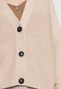 Beige gebreid vest met een V-hals ontwerp, vijf zwarte knopen aan de voorkant en een gestructureerde stof.