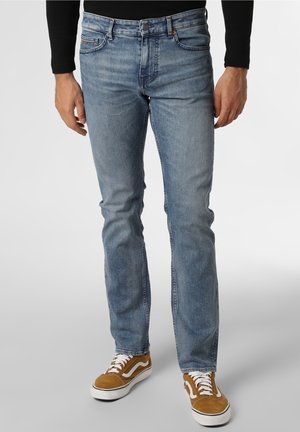 Homme portant un jean slim bleu clair et des baskets marron avec des lacets blancs, debout devant un fond uni.