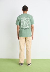Green cotton t-shirt with fish graphic print on the back. Paired with cream-colored, loose-fitting pants and black and white shoes.