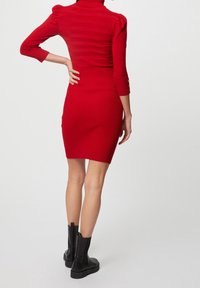 Rotes, geripptes Kleid mit tailliertem Schnitt, Rollkragen und puffärmeln; hat Knopfdetails an den Ärmeln. Schwarze Knöchelstiefel.