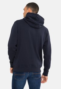 Felpa blu navy con una vestibilità comoda, tessuto testurizzato e cappuccio con cordino. Indossata con jeans blu, che mostrano polsini e orlo a costine.