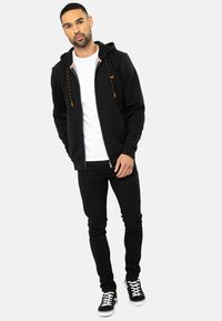 Sort hættetrøje med orange-detaljerede snore, lynlås lukning og sidelommer. Kombinationen er med sorte jeans og sorte sneakers med hvide såler.