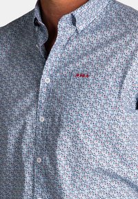 Lichtblauwe getailleerde shirt met een klein bloemenpatroon in groen en rood. Heeft een button-down kraag en een geborduurd logo op de borstzak.