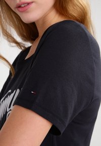 Camiseta de algodón negra con escote redondo y mangas cortas. Presenta un pequeño acento tricolor en la manga. Textura suave, diseño casual.