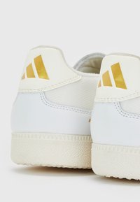 Baskets blanches avec une semelle en caoutchouc texturée, présentant des accents dorés et un matériau lisse au niveau du talon avec trois rayures diagonales.