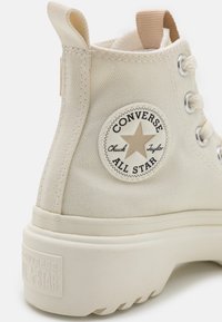 Białe trampki w fasonie za kostkę z okrągłą łatą z logo, metalowymi oczkami, teksturowanym materiałem i grubą gumową podeszwą.