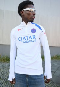 Hvid langærmet sportsjersey med halv-lynlås krave, blå detaljer, rødt Nike-logo og teksten "Qatar Airways" i blå. Tekstureret stof.