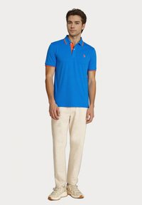 Blå polo skjorte med orange detaljer, korte ærmer og kravesnit. Kombineret med lyse beige bukser og brune sneakers.