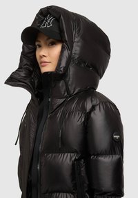 Musta, muhkea untuvatakki, jossa on iso huppu, vetoketjullinen etuosa, säädettävät nyörikahvat ja logopatch hihaosa.