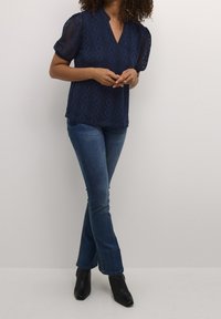 Mörkblå spetsblus med korta puffärmar, V-ringning och geometriskt mönster, kombinerad med mörkblå utsvängda jeans och svarta ankelboots.