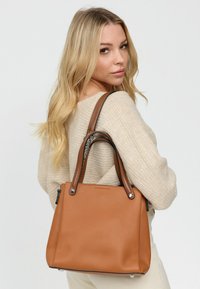 Tan gekleed lederen tote bag met dubbele hengsels, zilveren hardware accenten en een gestructureerde vorm. Rits-zak aan de zijkant zichtbaar.