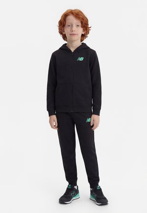 Kind mit roten Haaren steht in einem schwarzen New Balance Trainingsanzug und Sneakers vor einem einfarbigen weißen Hintergrund.