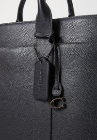 Struktureret tote bag i sort læder med tekstureret overflade, der har håndtag, et mærke med prægede logo og metal hardware detaljer.