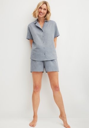 Lachende vrouw staat blootsvoets tegen een witte muur, gekleed in een lichtblauw korte mouwen overhemd met knopen en bijpassende korte broek.