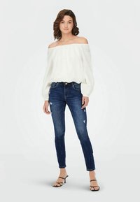 Balta off-the-shoulder bluzė su plačiomis rankovėmis derinama su tamsiai mėlynomis suplėšytomis lieknomis džinsomis. Modelis dėvi juodus šlepetes su dirželiais. Paprasta fonas.