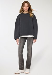 Felpa oversize grigio scuro con scollo rotondo, indossata sopra una T-shirt bianca, abbinata a jeans grigi aderenti e sneakers bianche.