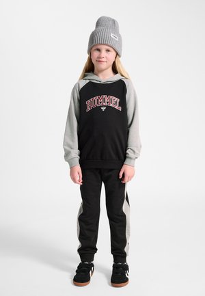 Junges Kind steht, trägt graue Strickmütze, schwarzen und grauen Hoodie mit der Aufschrift "HUMMEL", passende schwarze und graue Hose sowie schwarze Turnschuhe.