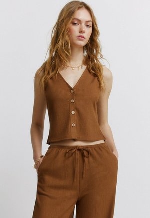 Kobieta z falowanymi włosami, ubrana w brązowy bezrękawnik zapinany na guziki oraz pasujące spodnie ze sznurkiem, stoi z rękami w kieszeniach.