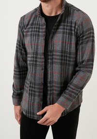 Camicia a quadri grigia con dettagli rossi, maniche lunghe, chiusura con bottoni e un tessuto morbido e texture. Indossata sopra una maglietta nera.