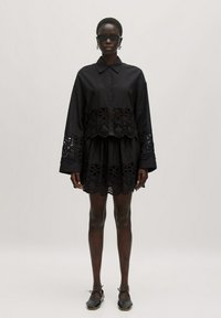 Schwarze Button-Down-Bluse mit langen Ärmeln und durchbrochenen Ösenmustern. Kombiniert mit einem passenden schwarzen Stufenkleid. Getragen mit dunkler Sonnenbrille.