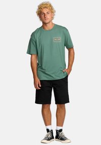 Groene katoenen t-shirt met korte mouwen en een borstafdruk, gecombineerd met zwarte shorts en hoge zwarte sneakers.