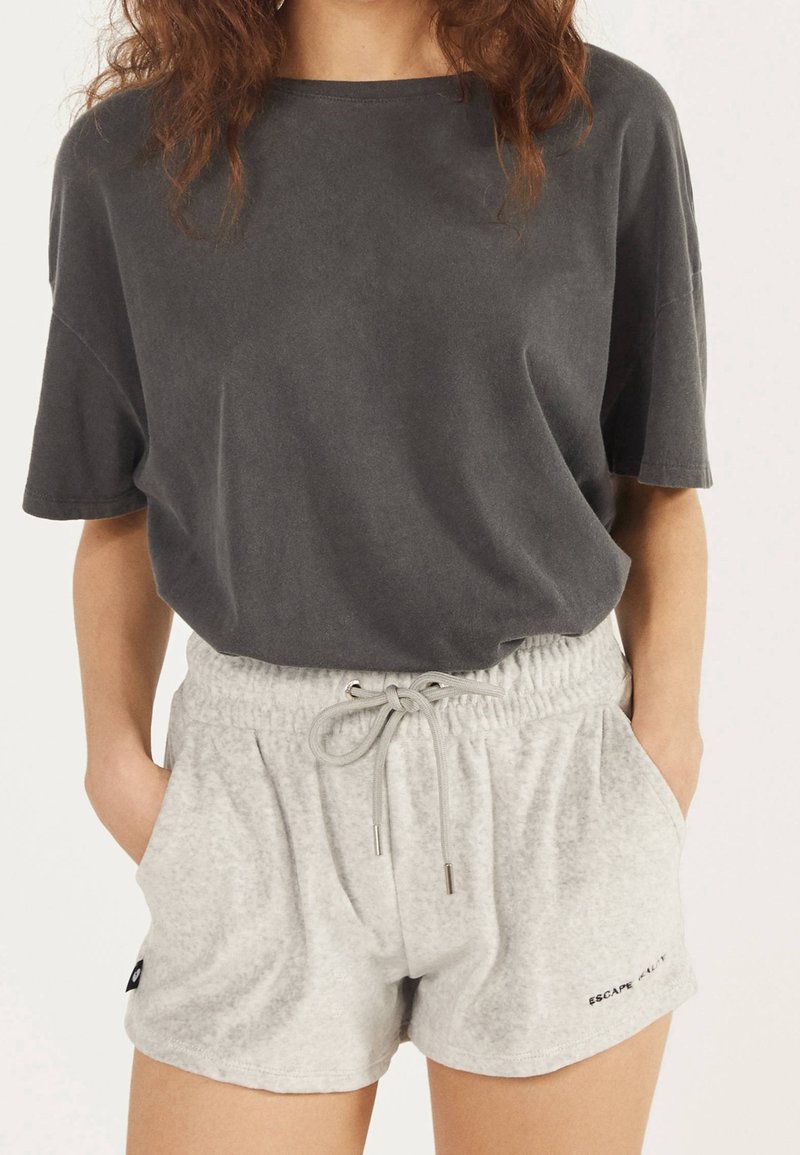 Femme portant un t-shirt ample à manches courtes gris foncé et un short gris clair à cordon avec poches, debout les mains dans les poches.