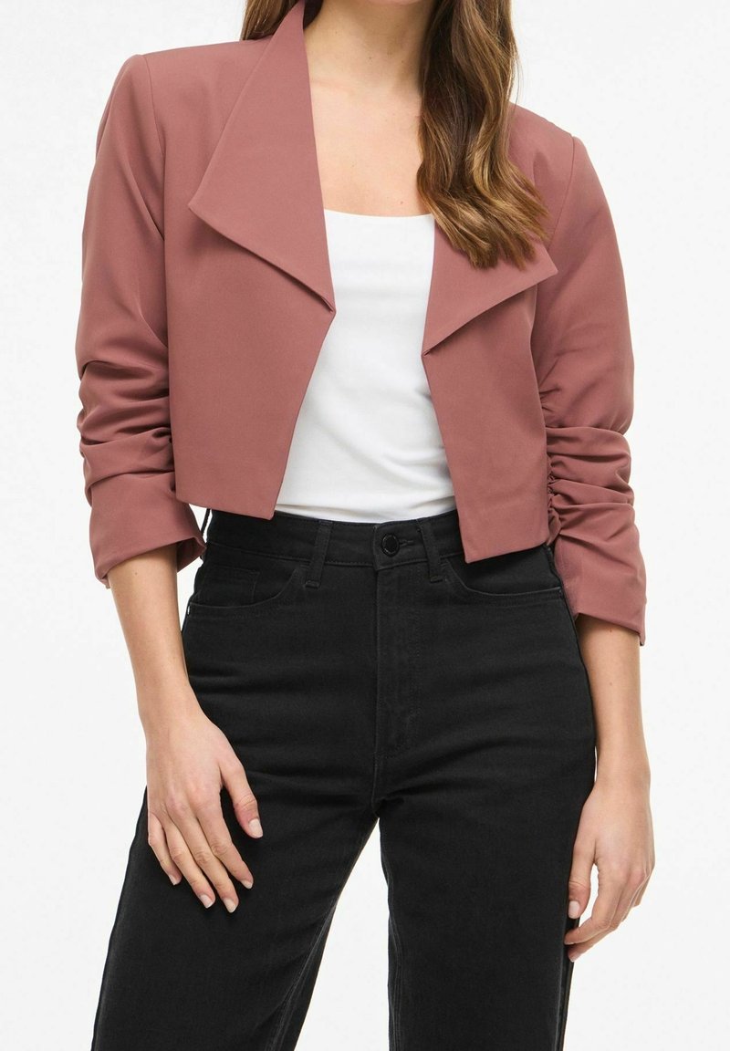 Femme portant une veste courte rose poudré sur un haut blanc avec un jean noir taille haute, debout les mains détendues le long du corps.