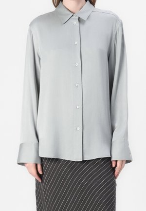 Femme portant une chemise gris clair à manches longues avec de larges poignets et une jupe foncée à fines rayures blanches diagonales.