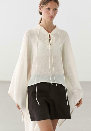 Vrouw draagt een losse, halfdoorzichtige witte poncho met lange striklinten over zwarte shorts, staand tegen een effen lichte achtergrond.