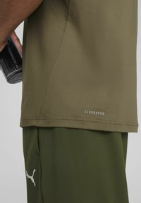 Camiseta verde que absorbe la humedad con un dobladillo redondeado. Presenta un logo "CLOUDSPUN" en gris claro cerca de la parte inferior. Tejido suave y ligero.
