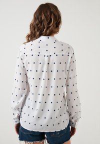 Femme aux cheveux bruns ondulés portant une chemise blanche à manches longues à pois bleus et un short en denim foncé déchiré, vue de dos.