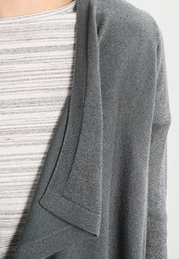 Světlý šedý kardigan s jemnou pletenou texturou, s volně splývajícím otevřeným předním dílem a ohrnutými manžetami, vrstvený přes pruhovaný šedobílý top.