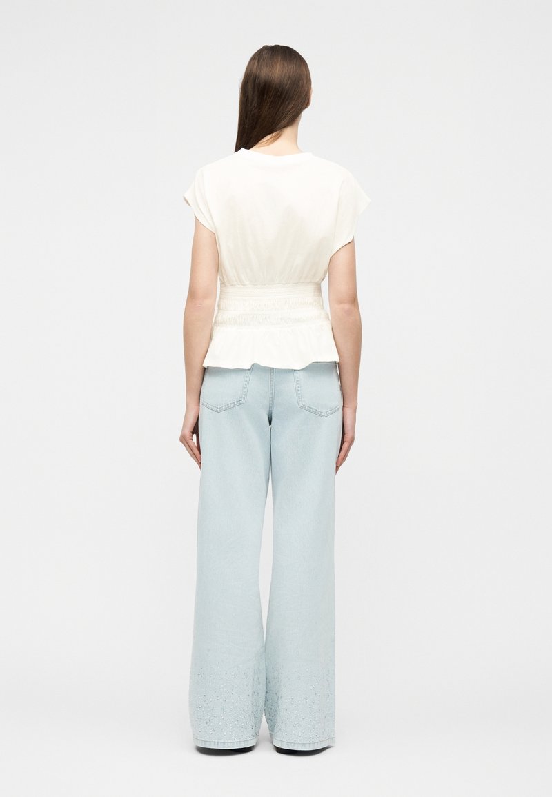 Femme aux longs cheveux bruns portant un top blanc à manches courtes et un jean large bleu clair, debout, de dos sur un fond uni.