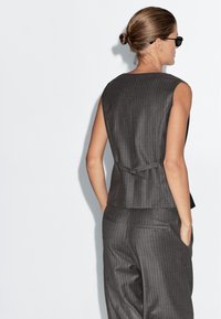 Kobieta z włosami związanymi w kok, nosząca czarne okulary i ciemnoszary garnitur bez rękawów w prążki, z rękami w kieszeniach, odwrócona plecami.