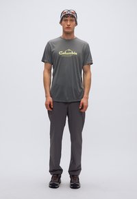 Γκρι κοντομάνικο πουκάμισο απορροφητικό της υγρασίας με κίτρινο λογότυπο «Columbia», σε συνδυασμό με γκρι ελαφριά παντελόνια και παπούτσια με αντίθεση, φορεμένα από μοντέλο.