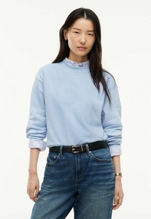 Kvinde med langt mørkt hår iført lyseblå sweatshirt over en stribet skjorte stoppet ned i blå jeans med sort bælte og guldfarvet ur.