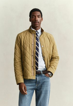 Homme portant une veste matelassée beige, une chemise blanche, une cravate rayée bleu marine, un jean bleu et une montre, debout avec une main dans la poche.