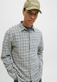 Chemise à manches longues à carreaux en vert clair, bleu et blanc avec un col boutonné; associée à une casquette beige ornée de texte brodé.