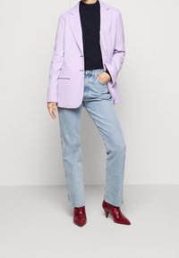 Blazer lavanda chiaro su una maglia nera, abbinato a jeans azzurri chiari e stivali rossi a punta. Tessuto morbido e vestibilità sartoriale.