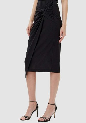 Gonna nera al ginocchio realizzata in tessuto elasticizzato, caratterizzata da un dettaglio annodato in vita e design drappeggiato, abbinata a tacchi neri con cinturini.
