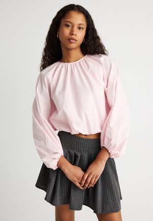 Junge Frau mit langem lockigem Haar, die eine hellrosa Bluse mit Puffärmeln und einen grauen, fein gestreiften Faltenrock trägt.