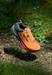 Chaussure de trail en orange et gris, présentant un dessus texturé, un système de laçage proéminent et une semelle robuste avec une adhérence multidirectionnelle.