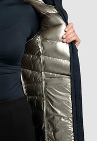 Izoliuota striukė, turinti tamsiai mėlyną išorinę pusę ir blizgantį metalinį sidabrinį pamušalą, demonstruojanti siuvin�ėtą paminkštinimą ir užtrauktuku užsegamą priekį.