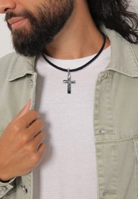 Collana nera intrecciata con ciondolo a croce in argento. Il ciondolo presenta un design testurizzato e pende al centro di una camicia bianca.