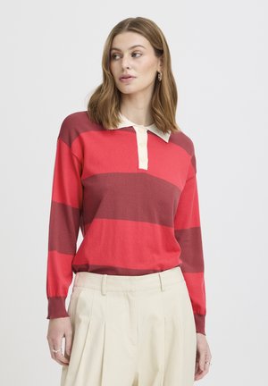 Kvinde med skulderlangt hår iført rød og mørkerød stribet langærmet poloshirt og cremefarvede højttaljede plisserede bukser.