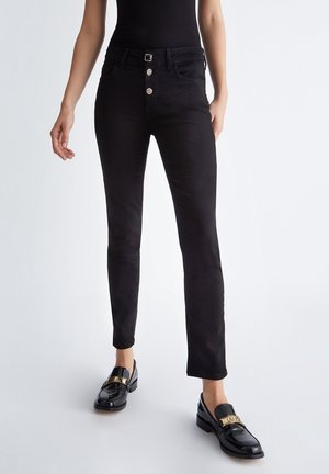 Woman wearing black skinny jeans with buttons and black shiny loafers with gold buckle details, standing against a light background.