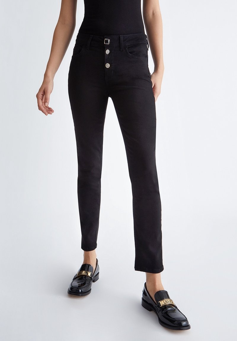 Femme portant un jean skinny noir avec des boutons et des mocassins noirs brillants avec des détails de boucle dorée, debout devant un fond clair.